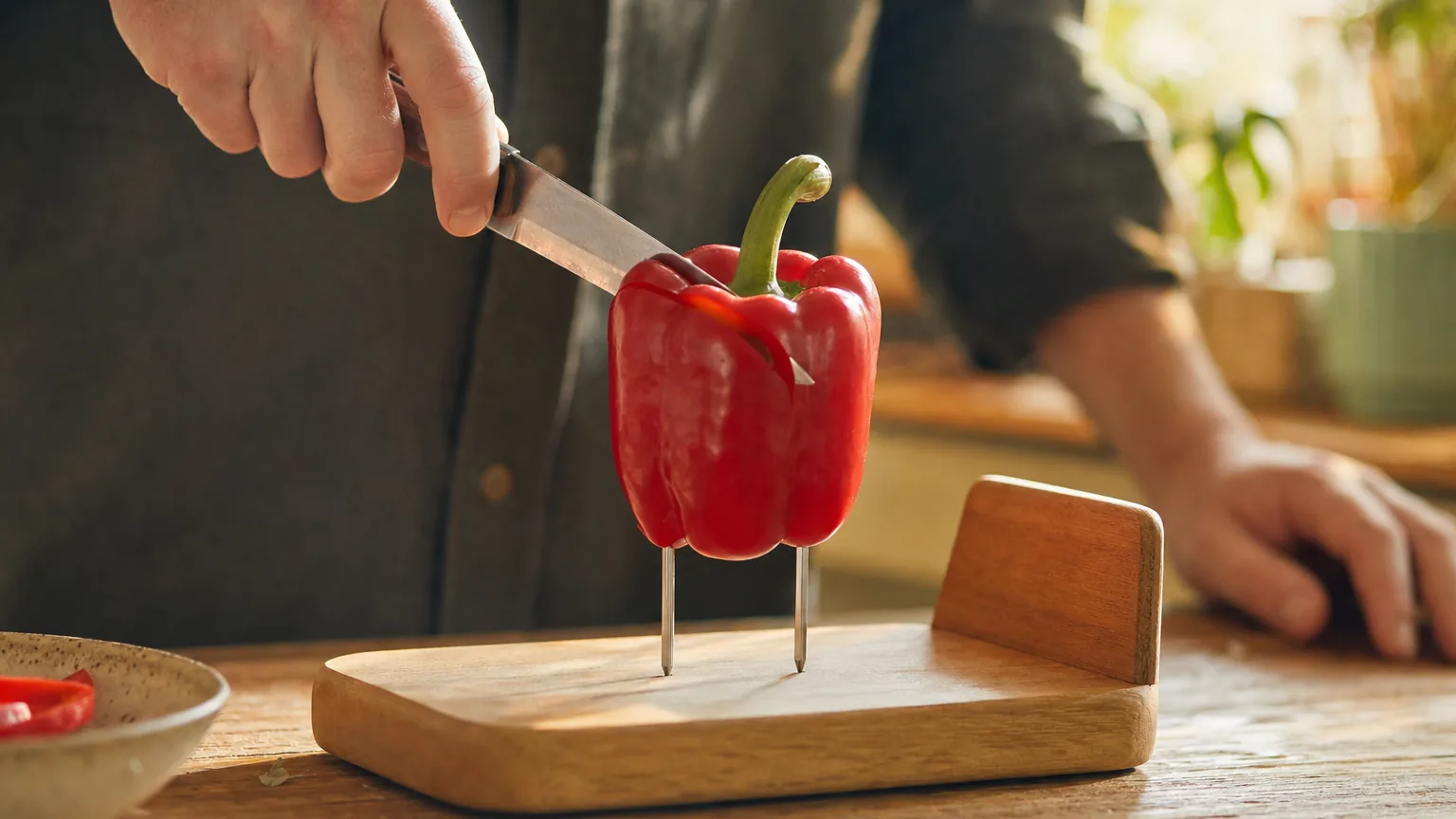 A hand slicing a red bell pepper held upright on the two spikes of a one-handed cutting board.
