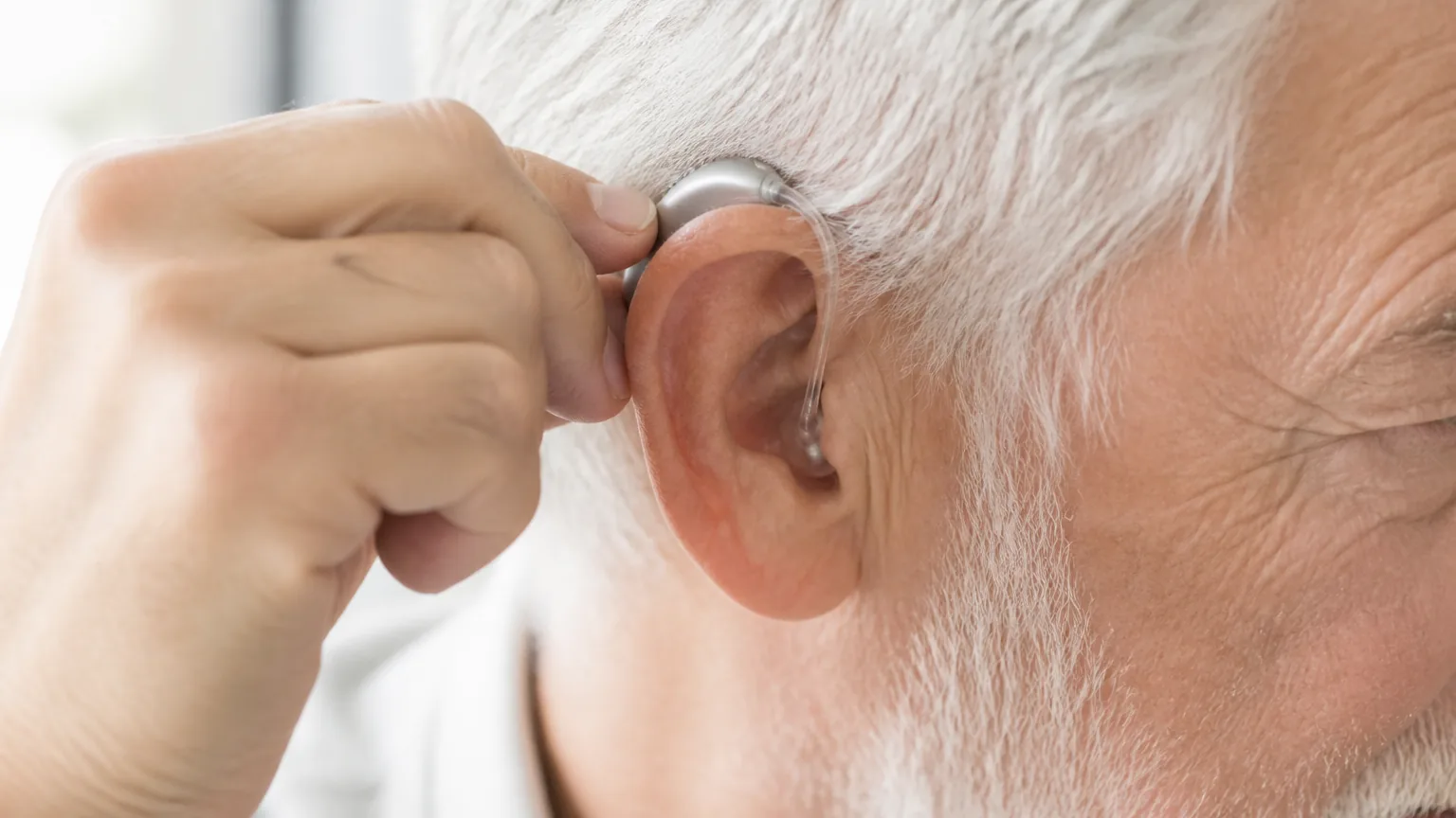 An older person placing a small modern behind-the-ear hearing aid into their ear.