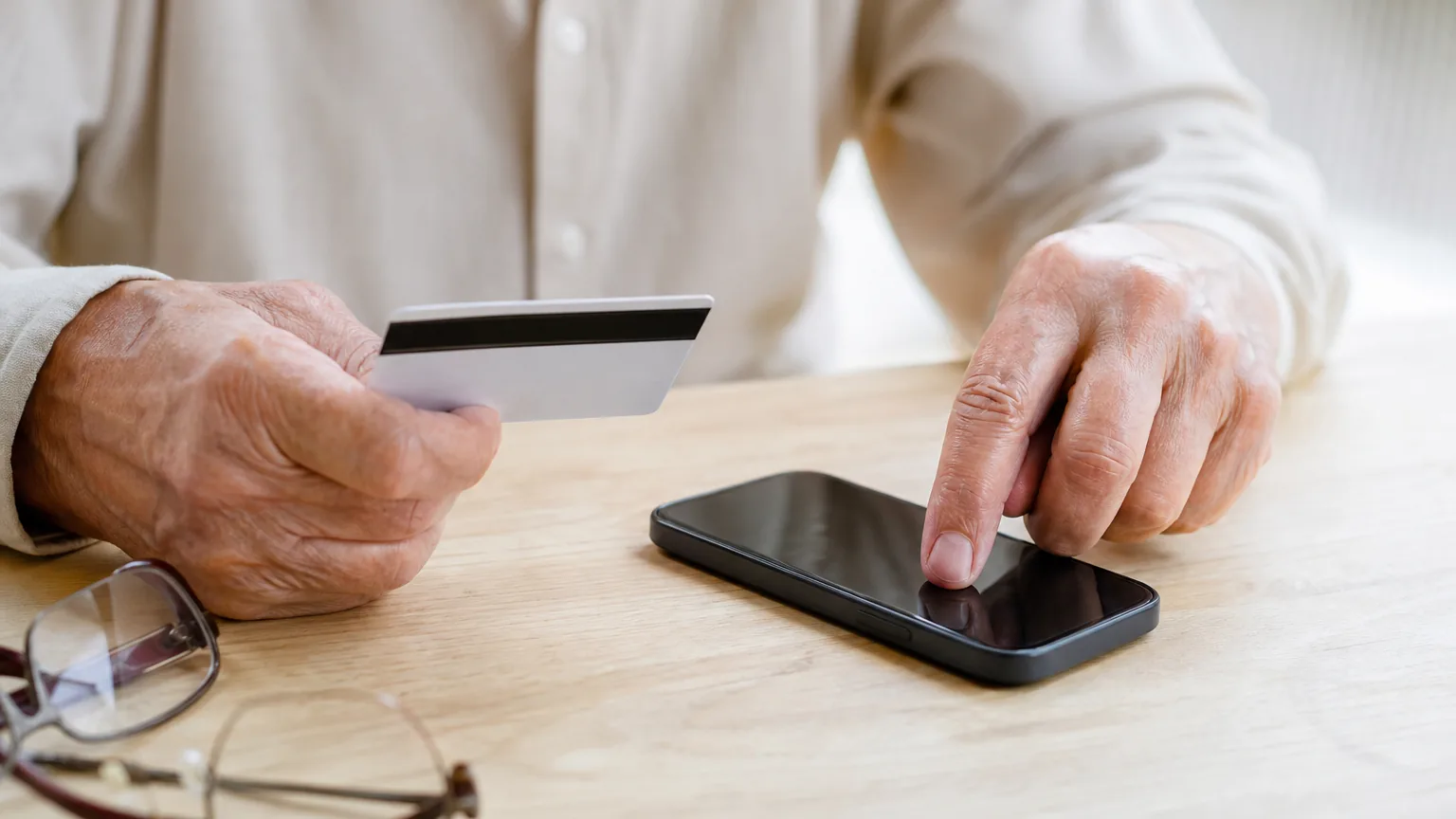 An older person holds a payment card turned to its back while dialing on a smartphone at a wooden table.